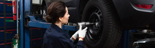 Vista lateral de la mujer que trabaja con llave de impacto y rueda de coche en el garaje, pancarta - foto de stock