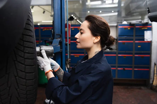 Side view of forewoman in uniform using impact wrench on wheel of car — Fotografia de Stock