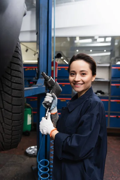 Smiling mechanic looking at camera and holding impact wrench near car in service — Stock Photo
