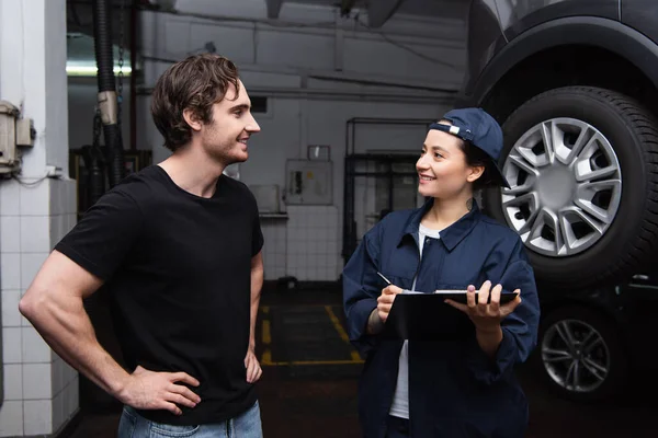 Positive driver standing near workwoman with clipboard near car in service — Stock Photo