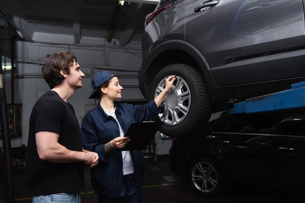 Side view of forewoman with clipboard pointing at car wheel near driver in garage — Stock Photo