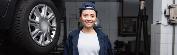 Positive mechanic in cap looking at camera near car in service, banner — Stock Photo