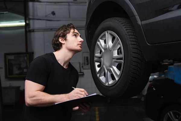 Mechanic writing on clipboard and looking at car in service — стоковое фото