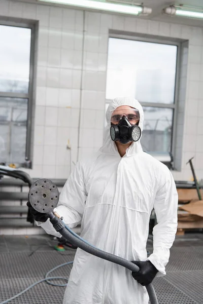 Workman in hazmat suit and respirator holding car polisher in garage — Stock Photo