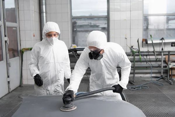 Workman in hazmat suit and respirator polishing auto near colleague in garage — Foto stock