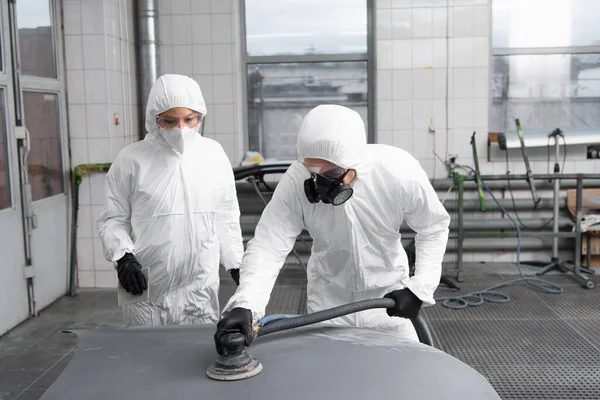 Workman in hazmat suit and respirator polishing auto near colleague in garage — стоковое фото