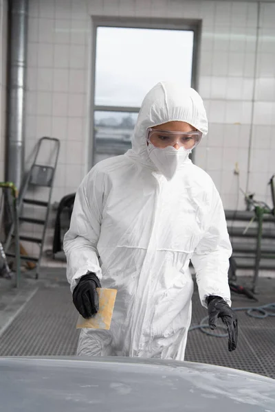 Workwoman in hazmat suit holding sandpaper near car in garage — Fotografia de Stock