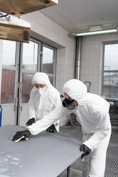 Workman in hazmat suit working with sandpaper and car hood near colleague in garage — Foto stock