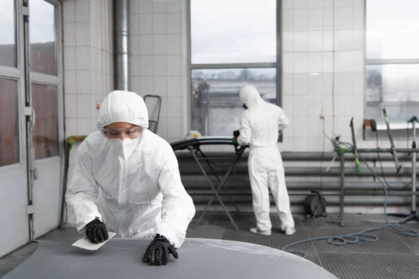 Workwoman in goggles and hazmat suit holding sandpaper near car hood in garage — Stock Photo