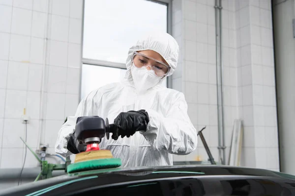 Workwoman in hazmat suit and protective mask polishing car in service — стоковое фото