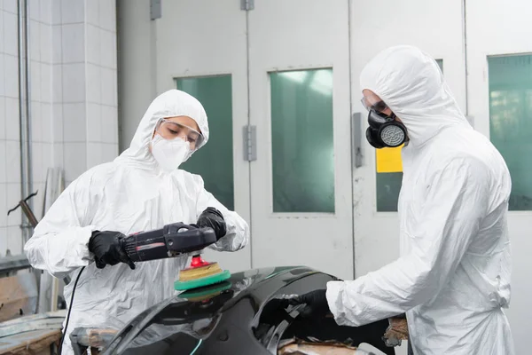 Trabajadora en traje de materiales peligrosos pulido pieza de coche cerca de colega en respirador en servicio - foto de stock