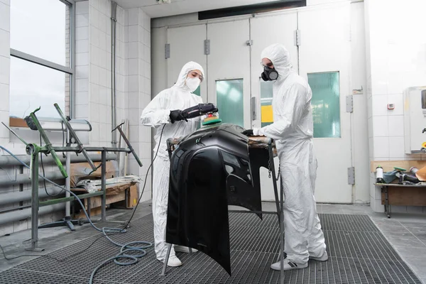 Workwoman polishing car part near colleague in hazmat suit in garage — стоковое фото