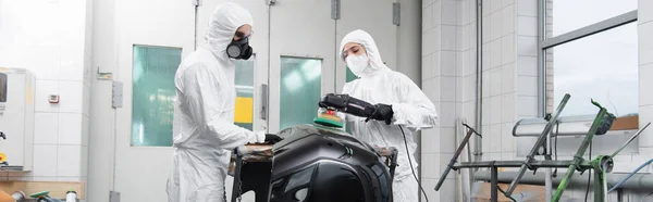 Trabajador en traje de materiales peligrosos que sostiene la pieza del coche cerca de colega con pulidor en servicio, pancarta - foto de stock