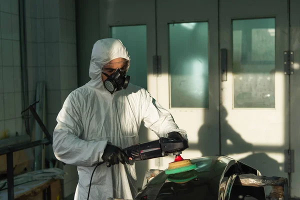 Workman in protective suit and respirator polishing car part in garage — Foto stock