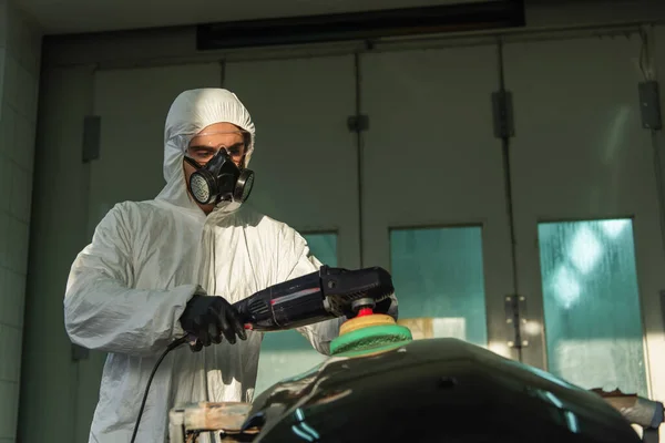 Workman in hazmat suit and respirator polishing auto part in service — стоковое фото