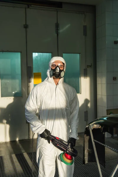 Workman in respirator and hazmat suit holding car polisher and looking at camera in garage — Foto stock
