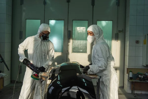 Workman in protective suit polishing car part near colleague in service — Stock Photo