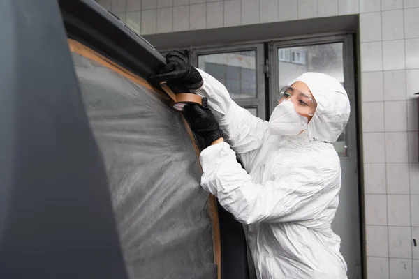 Trabajadora en traje de materiales peligrosos aplicando cinta adhesiva en el coche en el garaje - foto de stock
