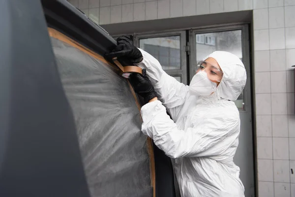 Workwoman em terno hazmat aplicando fita no carro na garagem — Fotografia de Stock