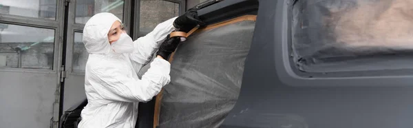 Workwoman in protective suit and gloves applying tape on car in garage, banner — Stock Photo