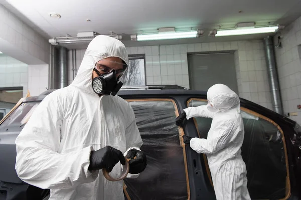 Trabajador en traje de materiales peligrosos sujetando cinta cerca de colega y coche en el garaje - foto de stock