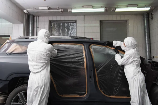 Workwoman applying tape on car with cellophane near colleague in service — стоковое фото