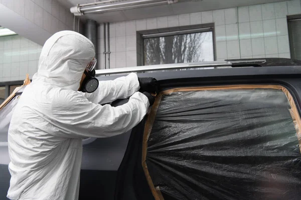 Ouvrier en respirateur et combinaison Hazmat appliquant du ruban adhésif sur le cellophane sur l'automobile en service — Photo de stock