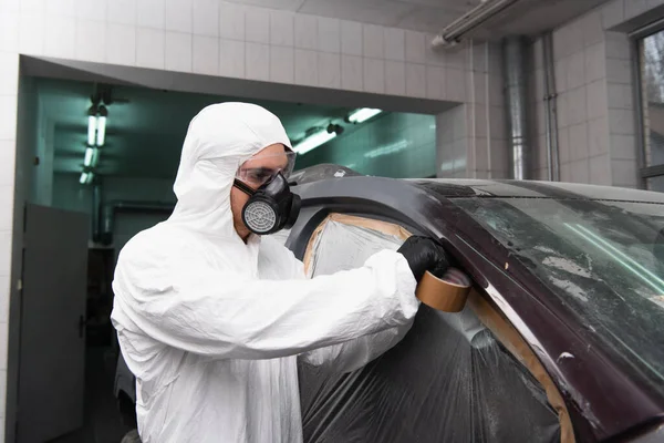 Travailleur dans un respirateur et lunettes appliquer du ruban adhésif sur le cellophane tout en travaillant dans le service de voiture — Photo de stock