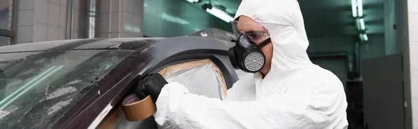 Young workman in hazmat suit applying tape on car with cellophane in garage, banner — стоковое фото