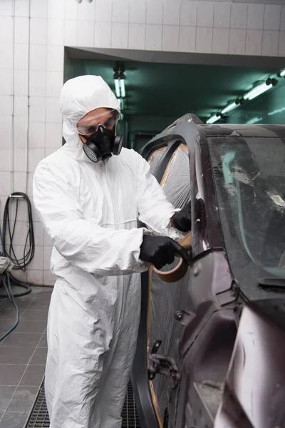 Workman in hazmat suit applying duct tape on cellophane in car in garage — Stockfoto