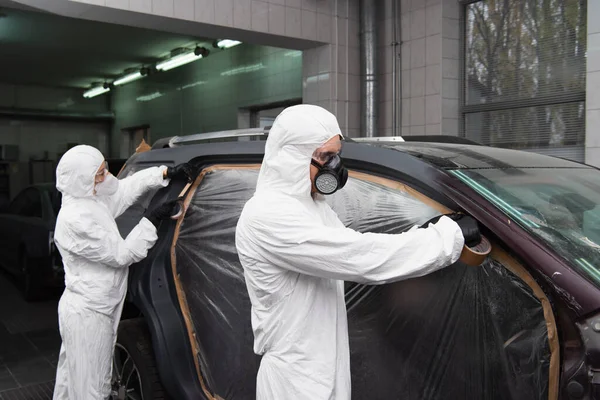 Foreman in hazmat suit applying duct tape and cellophane on car near colleague in service — Stock Photo