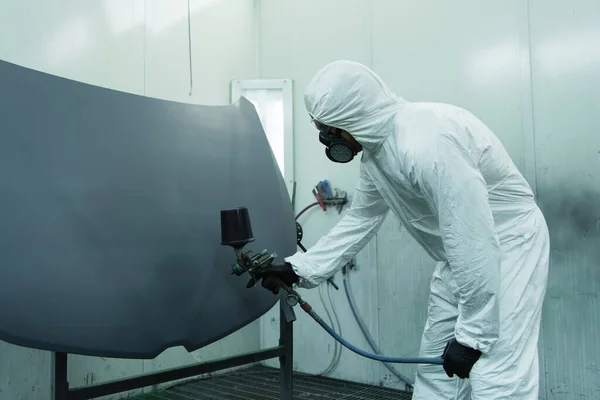 Workman in protective suit spraying varnish on car part in garage — Stock Photo