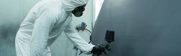 Workman in hazmat suit spraying varnish on car part in garage, banner — Foto stock