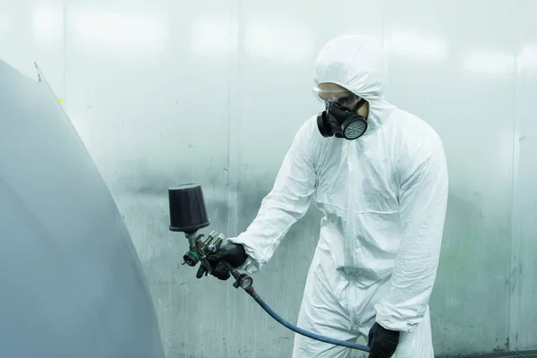 Workman in hazmat suit and respirator using aerograph on car part in garage — Stockfoto