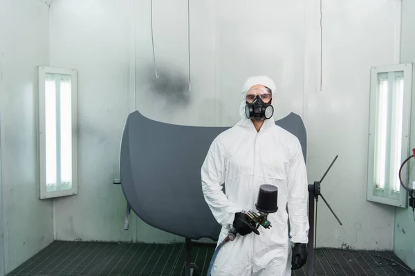 Workman in protective suit and respirator holding aerograph and looking at camera in garage — стоковое фото