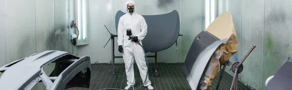 Young workman in hazmat suit holding airbrush near car parts in service, banner — Stockfoto