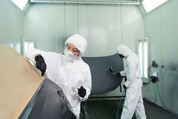 Femme de travail en lunettes et combinaison de protection travaillant avec la pièce de voiture près de collègue avec aérographe dans le garage — Photo de stock