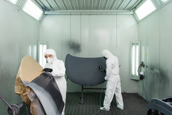 Young workwoman in hazmat suit and protective mask standing near car part while colleague using airbrush in garage — Stockfoto