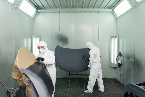 Young workwoman in hazmat suit and protective mask standing near car part while colleague using airbrush in garage — Stock Photo