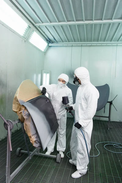 Workman in hazmat suit holding airbrush near colleague pointing with finger at car part in service — Stockfoto