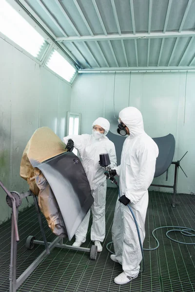 Workman in hazmat suit holding airbrush near colleague pointing with finger at car part in service — Stock Photo