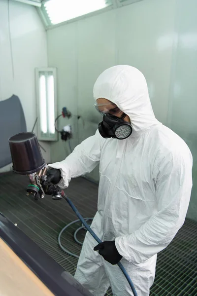 Workman in hazmat suit coloring part of car with airbrush in garage — Stockfoto