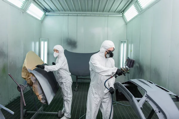 Workman coloring car part near colleague in hazmat suit in garage — Stock Photo