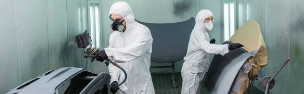 Trabalhador em hazmat terno colorir parte do carro com aerógrafo perto colega em máscara protetora na garagem, banner — Fotografia de Stock