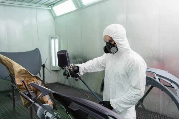 Young workman holding airbrush while coloring car part in garage — Stockfoto