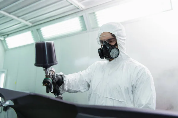 Workman in hazmat suit and respirator coloring blurred car part in service — Stock Photo