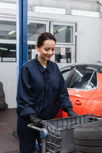 Smiling workwoman standing near tire on cart in garage — Stock Photo