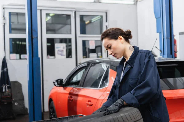 Femme de travail tatouée avec des gants tenant un pneu près d'une voiture floue dans un garage — Photo de stock