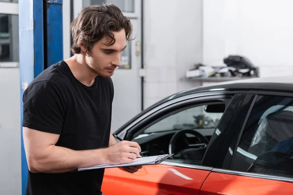 Joven mecánico escribiendo en el portapapeles cerca de coche en el garaje - foto de stock