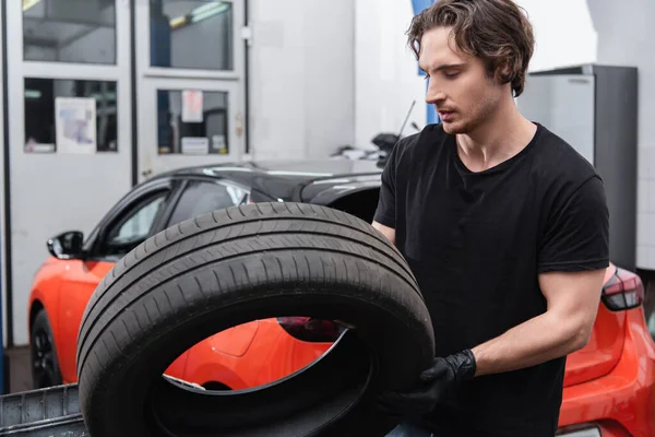 Young workman in gloves holding tire near blurred car in service — стоковое фото